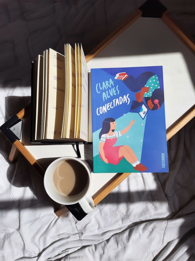 livro Conectadas da Clara Alves em uma mesinha de madeira do lado de uma caneca de café com leite