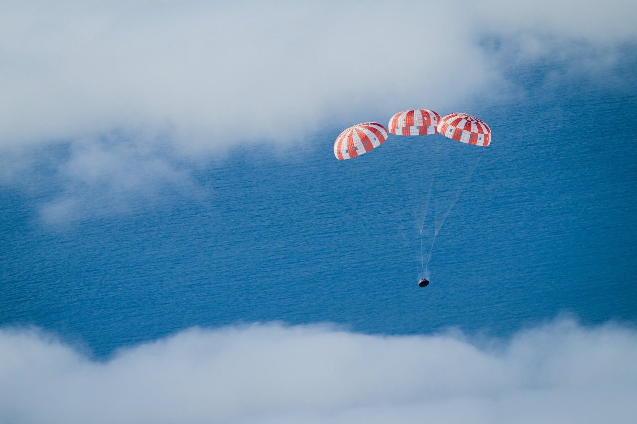 The Orion spacecraft parachutes down toward splashdown. The three main parachutes are patterned with white and red stripes. Orion stands out against a backdrop of a bright blue ocean. Steam comes off the crew module as it passes through the cloud layer. Credit: NASA