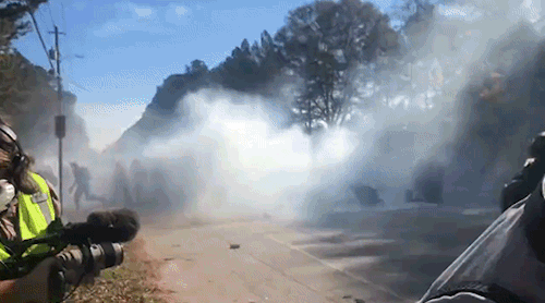 [GIF panning from a large cloud of tear gas, inside which silhouettes of many people are visible, to a crowd of police in full militarized tactical gear, carrying either clear plastic riot shields or automatic weapons, retreating away from the cloud. As they retreat, one of them calls out a warning about the gas and yells for them to "turn around"]