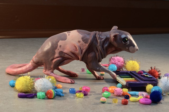 A photo of a hand-sized painted clay figurine of Neil the hairless rat, playing a small purple synthesizer. The photo is taken slightly from above, showing Neil\'s left side.  The knobs, wheels and black keys of the synth are in the colors of the rainbow. The figurine has its right forepaw raised, resting on the keys.  The figurine sits on a grey vinyl floor, and is surrounded by colorful glittery pompoms and plastic beads in a variety of shapes laid out around it. Some of the beads are shaped like stars and musical notes.