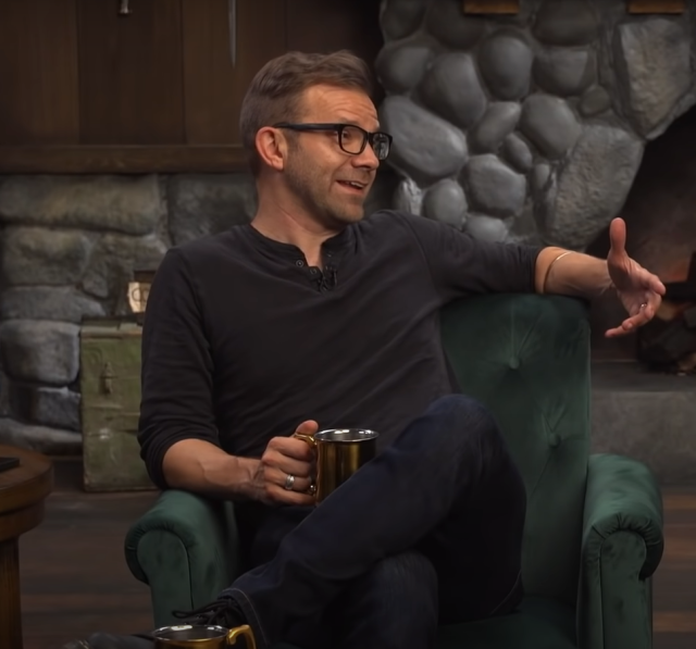 Liam has on a black henley, dark wash jeans and black boots along with black plastic frame glasses. He is speaking to Sam who is to his left and is gesturing slightly with his left hand, snake armlet just barely visible. 