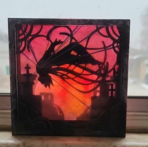 A softer light behind the craft, placed on a windowsill. The color of the red sky is deeper and less fiery, but more saturated. The silhouettes of the figure, the rooftops, and the frame are all much darker and the colors less distinct so the contrast effect is much higher. The furthest back rooftops appear faded into atmosphere and indistinct. The bright blue lines radiating from the center of the chest are much darker and appear black.