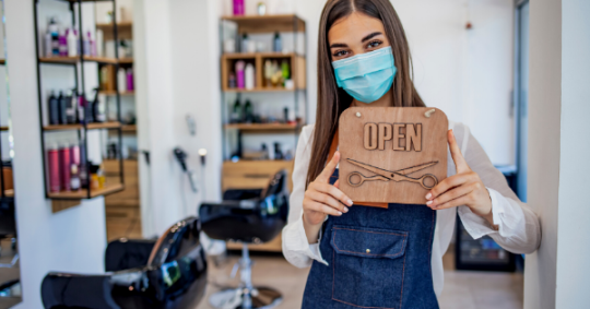 Image shows a young woman, presumably the owner of a small hair salon, with a mask on and holding a "Open" sign.