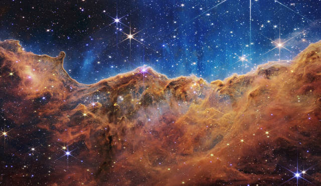 An undulating, translucent star-forming region in the Carina Nebula, hued in ambers and blues. Foreground stars with diffraction spikes can be seen, as can a speckling of background points of light through the cloudy nebula. Credit: NASA, ESA, CSA, and STScI