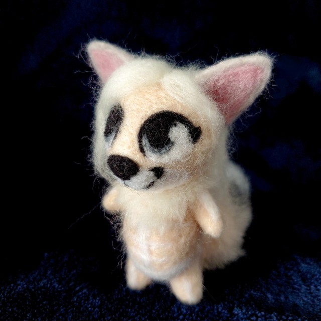 A photo of a needle felted sculpture of a fox-like creature named Menoes. She is peach colored with long, silver hair and white markings that look like bones. She is against a dark blue background.