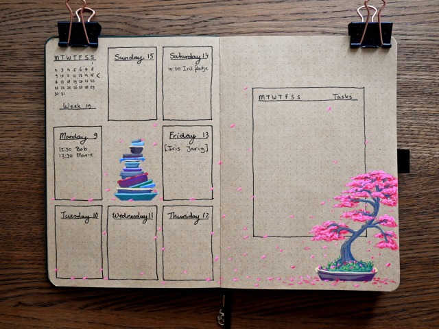 A photo of a bullet journal with pages of light brown kraft paper. On the left page a weekly layout is drawn, with the days of the week in black fineliner boxes, and a small overview of the days of the month in the top left corner. It is week 19. In the middle of the page is a drawing of a stack of different coloured and sized bonsai pots and containers. On the right page a 'rolling weekly' task list is drawn in the middle of the page. In the right bottom corner is a drawing of a large bonsai tree with pink leaves. Some of the pink leaves are drawn all over both pages, as if blown by wind across the pages and gathering near the bottom. 