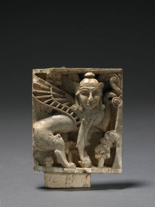 Ivory plaque carved in relief. Human headed winged sphinx wearing conical cap. One forepaw on a lotu