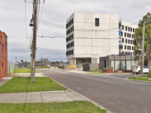 Derby Road Sunshine looking east in 2023. Th 6 storey building is complete and is made of concrete with windows that have a shade on them, there are still signs of construction in the street. The sky has soft cloud broken by patches of blue. The CBD skyline is visible on the horizon.