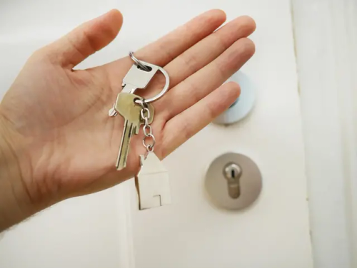 Hand holding two keys. A house figure is attached to the keychain. Background is a white door.