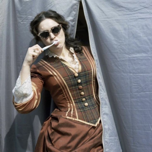 Danielle galligan peeking out of a black tent and brushing her teeth while she wears sunglasses and her orange and red season two costume 