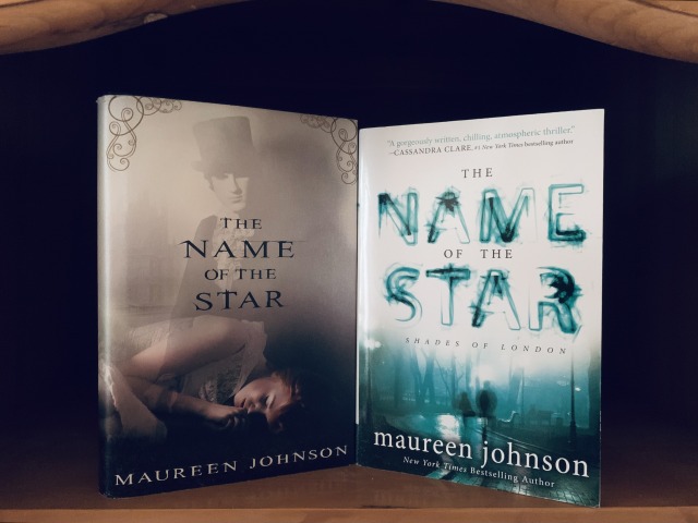 Shown are two editions of THE NAME OF THE STAR on a wood bookshelf. The cover art of the hardback edition shows a man in a top hat lurking in the shadows behind a sleeping young woman. The cover art of the softcover edition shows two shadowy figures walking on a cobblestone lane in the darkness. Photo by AHS.