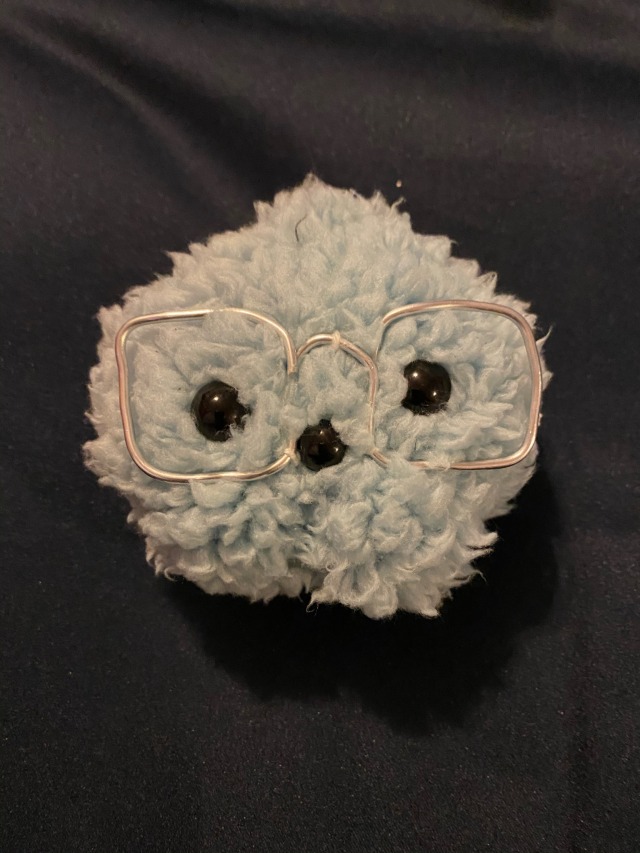 A small star shaped plush in blue. It has wire glasses, shiny black eyes, and a amall black mouth. It is made of fluffy faux fur fabric.