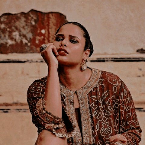 a plus size, dusky skinned indian woman sitting against a pale orange background. she has black hair in a knot, dark kohl-lined eyes and wears ornate jewellery and an embroidered russet kurta.