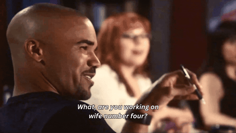 Derek Morgan, a man with a shaved head wearing a dark shirt, smiles and speaks. He is fidgeting with a pen. Out-of-focus, Penelope Garcia, a redhaired woman in glasses smiles with her mouth open. White subtitles read: "What, are you working on wife number four?"