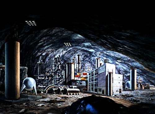 GIF 4: scenic view of a large cave. the image is panning from left to right. the cave is filled with equipment and larger building structures, for example what looks like a silo on the left, and a 4 story building in the center.