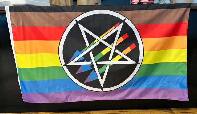 Rainbow Pride Flag with black and brown stripes at the top, a pentagram antifascist logo in the center. The arrows of the antifascist logo are also rainbow colored.
