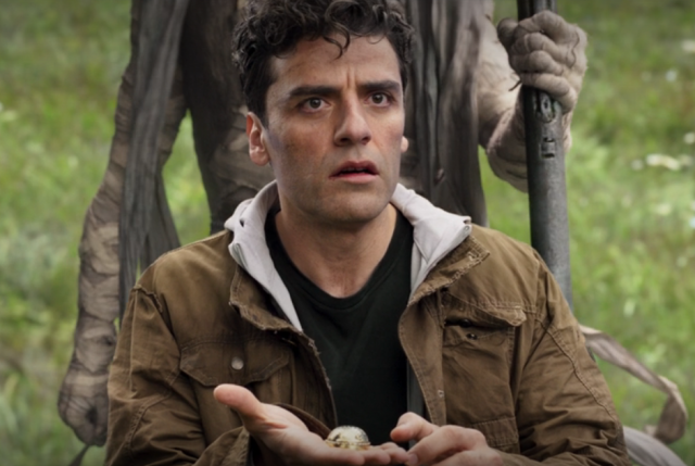 Screenshot showing Oscar Isaac looking confused in the foreground holding the same scarab as above, as a walking bandaged mummy moves ominously closer to him in the background. Only the mummy's torso and arms are visible, and they are using a walking stick.