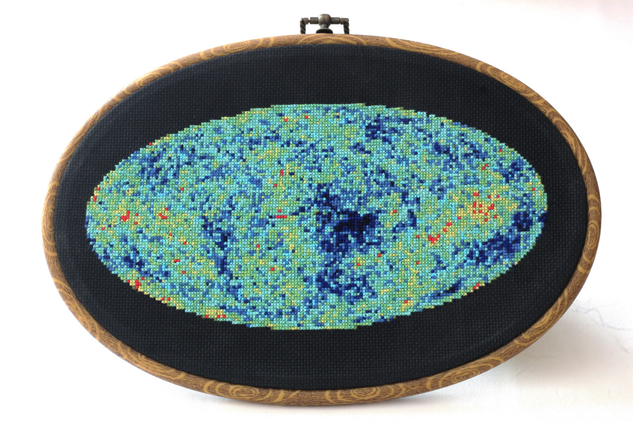 This image shows an embroidery design based on the cosmic microwave background, created by Jessica Campbell, who runs Astrostitches. Inside a tan wooden frame, a ccolorful oval is stitched onto a black background in shades of blue, green, yellow, and a little bit of red. Credit: Jessica Campbell/Astrostitches