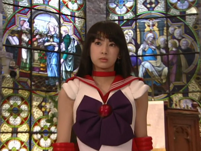 Sailor Mars (played by Keiko Kitagawa) looking disgruntled, standing in front of another pulpit and a wall of stained glass mirrors showing scenes from the bible, including the virgin Mary.