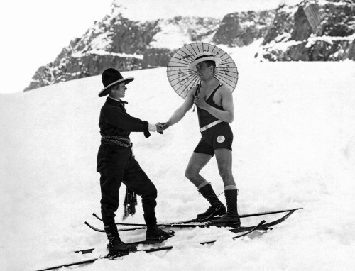 Adam Glickman - A woman skier on the mountain slope wearing a sash and western hat shakes hands with a man on skis in a one piece bathing suit and carrying a parasol, early to mid 1920’s.