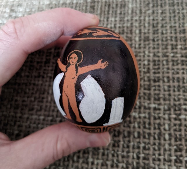 An egg that has been drawn on to resemble an ancient Greek red figure vase depicting Helen, small but fully formed, standing with her arms raised between two halves of an egg shell. This is from an angle to the right.