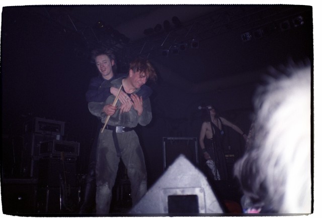 Blixa Bargeld hugging FM Einheit from behind. Blixa seems to have a cigarette in his right hand; Mufti has drumsticks in his. They're both smiling with their eyes closed. Alexander Hacke is standing in the background, to the right. There's the back of someone's head in the bottom-right corner.