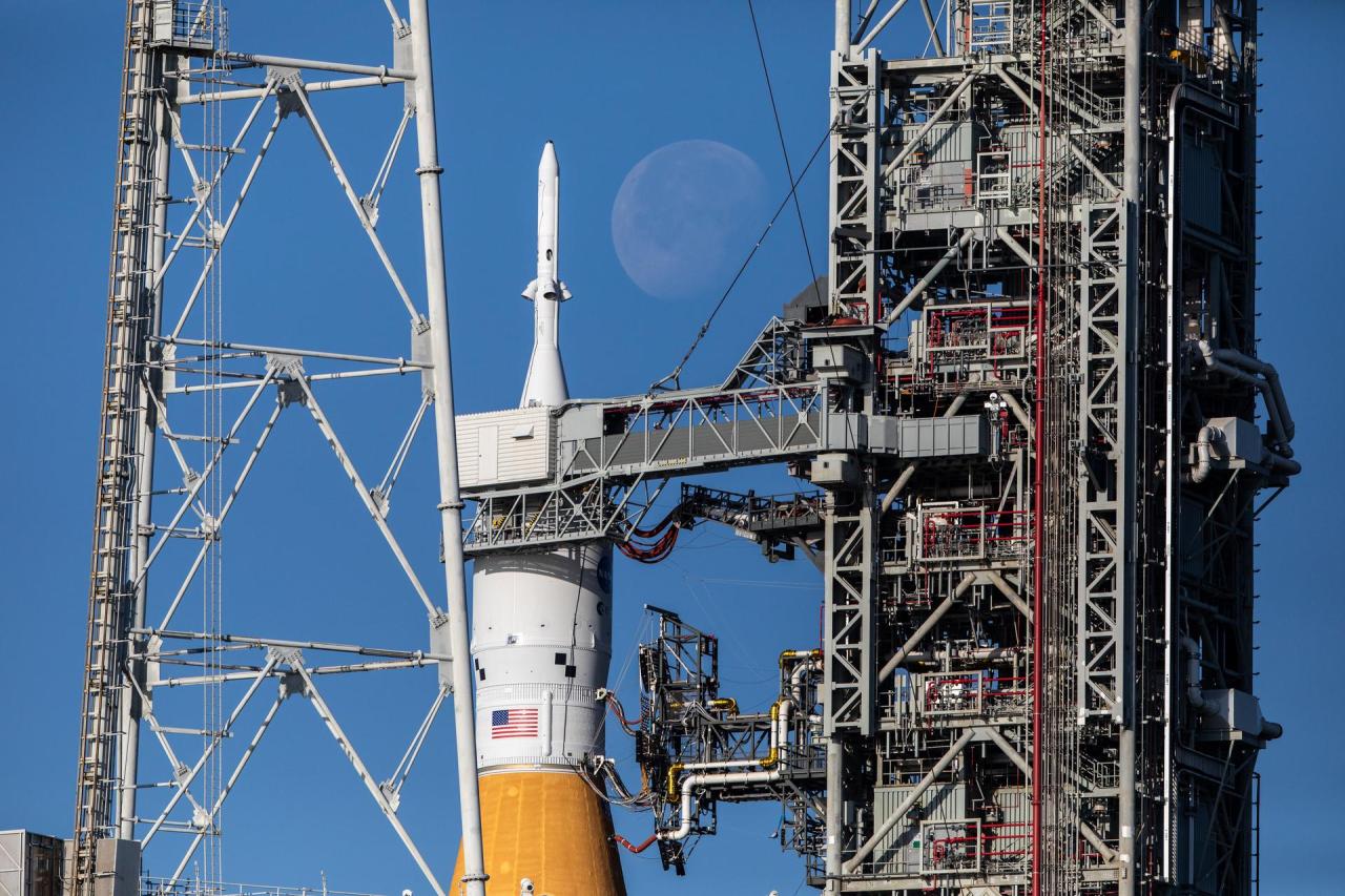 From left to right: A grey hollow pyramid-shaped lightning tower, the white Orion spacecraft and the top of the Space Launch System (SLS) rocket in orange, the Moon in faint white and gray, the Mobile Launcher with many pipes and levels in gray and red. The background is blue skies. Credit: NASA/Ben Smegelsky