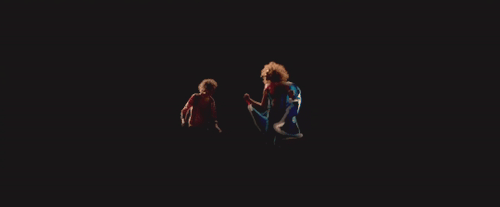 Both the young and present Danny are seen from the side in a pitch black space. They're facing each other and swinging their arms in dance.