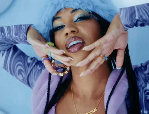 A light-skin black woman with freckles and her black hair in braids tilts her head up and looks into the camera, her hands caressing her face. She wears a light blue fur hat, blue eyeshadow and eyeliner, false eyelashes, lip gloss, multicolour rings, gold bamboo earrings, a gold nameplate that reads sagattarius, and purple, swirl-patterned cardigan with a purple, fur collar.