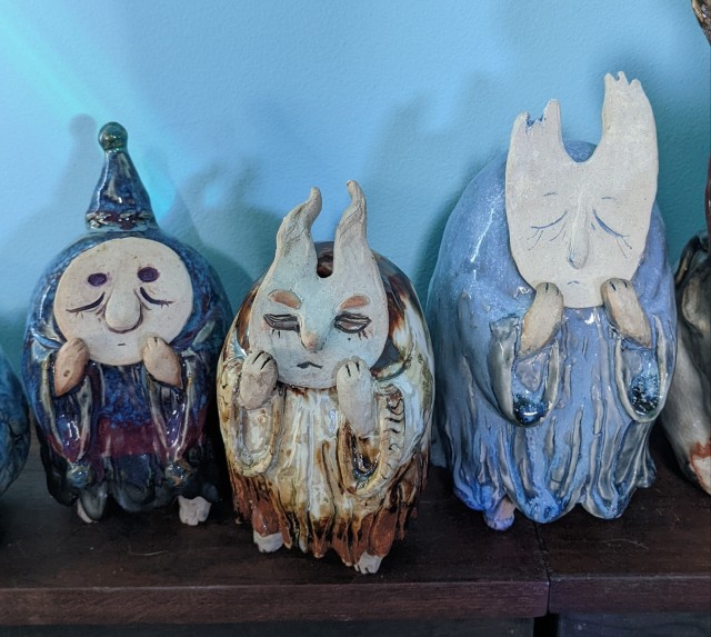 Three ceramic sculptures of little robed creatures standing on two feet clutching their faces. One has a pointy hat and a round, sleepy face. Their robe is blue and purple and black. One has a horned face, orange eyebrows, and a pointed nose. They are glazed in copper and cream. The last sculpture is the blue guy from the rest of the post.