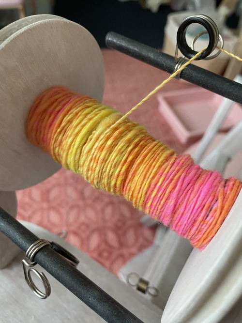 A wooden spinning wheel bobbin mounted on a spinning wheel. It’s in the process of being filled up with a neon yarn that is orange, yellow, and pink.