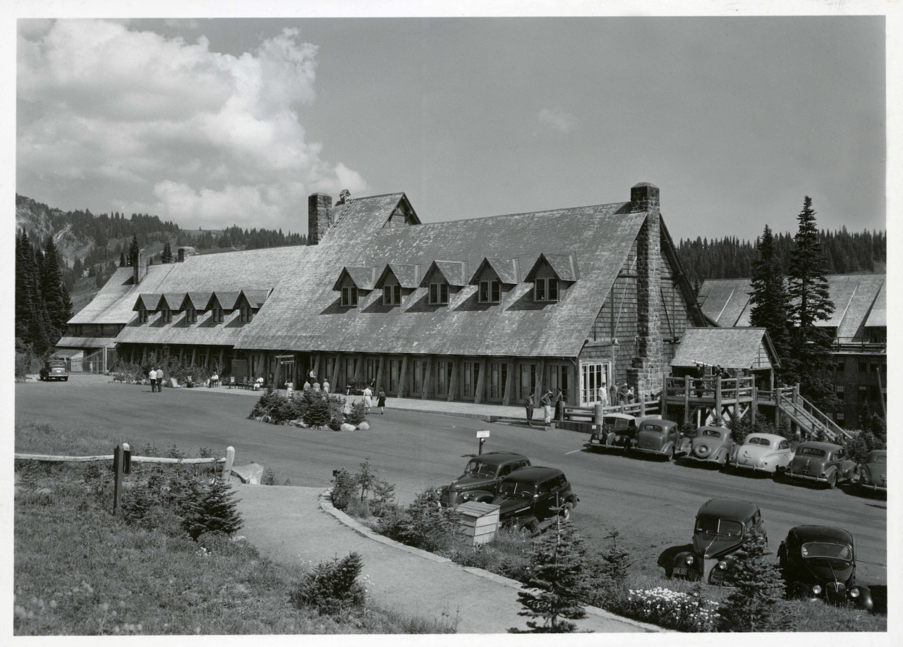 Black and white historic photo of a large wood building with a steeply angled shingle roof with gable windows and a covered walkway in front of the building.