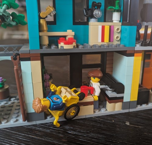 [Second image description: a photo of a Lego™ model coffee shop with the wheelchair-using figure on its back, awkwardly jammed against the coffee shop table. description ends]