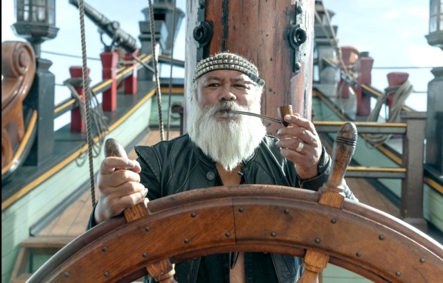 an image of dave fane portraying the character fang on our flag means death. he is standing behind the wheel/helm of a ship, puffing on a long stemmed pipe