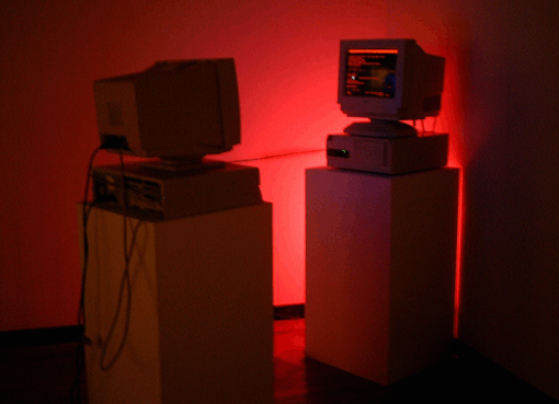 Two computers each on separate pillars, facing each other. A taut cable connects them. The scene is lit in red and the screen visible to the viewer displays scrambled, red text.