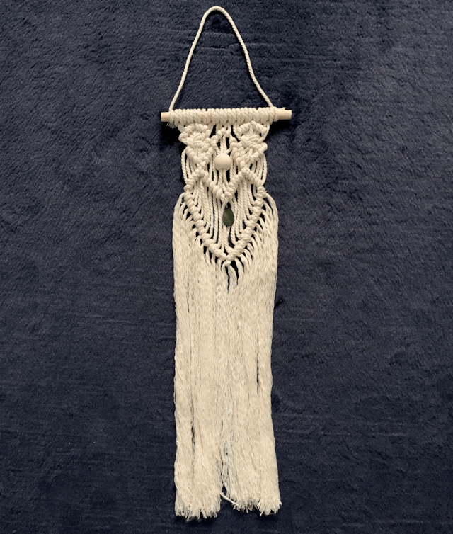 Photo of a white macrame wall hanging on a small wooden dowel, against a navy blue background. The macrame hanging has two diamond shapes at the top and a large heart shape below, with a white wooden bead above the heart and a green teardrop jewel within the middle of the heart shape. Underneath the heart shape, the cord is knotted and unraveled in long strands.