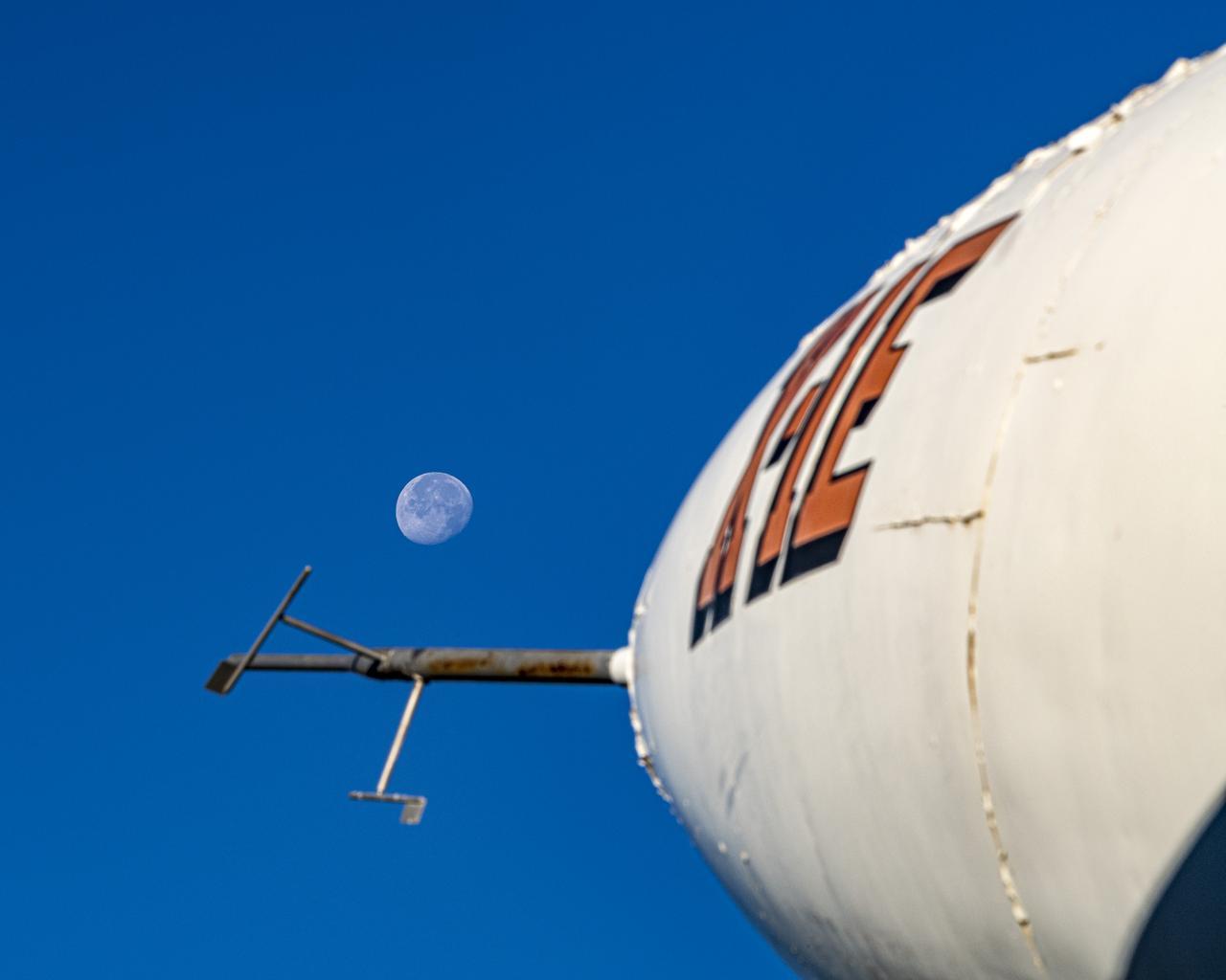 The X-1E aircraft dominates the foreground of this photo. It is white, with its designation written on it in big orange letters. The Moon is in the background, lined up with the nose of the airplane.