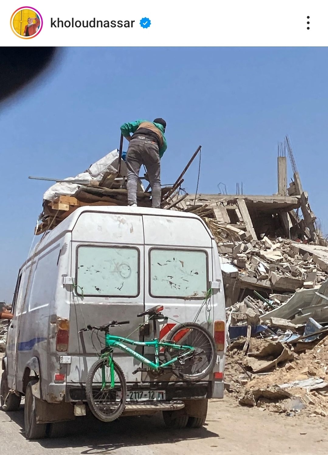 A van driving down a dusty road. Rubble of bombed buildings line the road. There's a bicycle hitched to the back of the van.