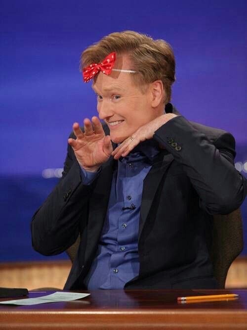 Conan O'Brien behind his desk in a blue shirt and black jacket. He is wearing a red and white polka dot bow on his forehead and smiling coyly with his hands under his chin.