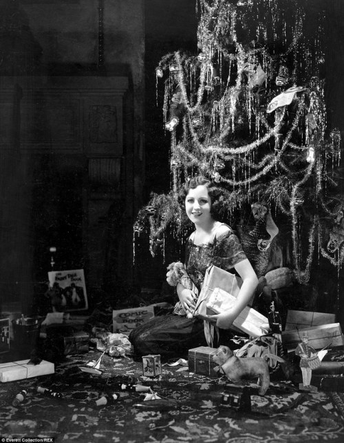 A woman sits at the foot of a tinsel-covered Christmas tree surrounded by string-tied presents inclu