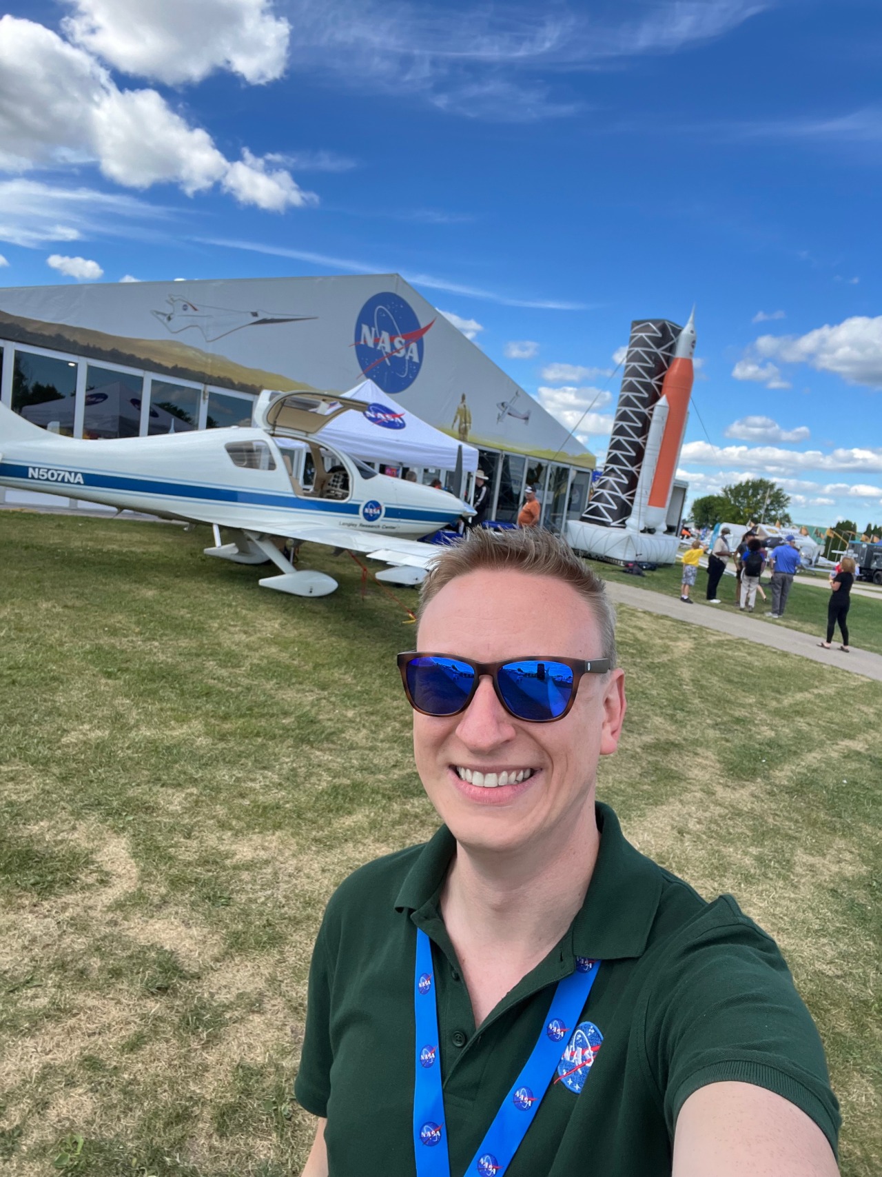 A man in a NASA polo shirt and lanyard takes a selfie in front of a white and blue emblazoned NASA aircraft and a large building with the red white and blue NASA logo imprinted on it.