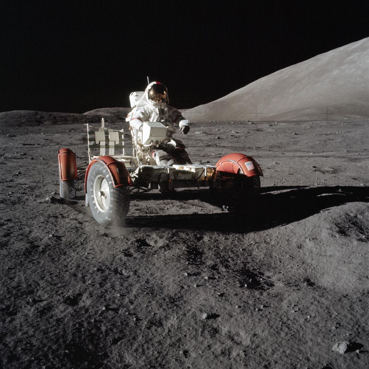 Astronaut Gene Cernan wears a bulky white space suit with a gold visor. He is sitting in the Lunar Roving Vehicle (LRV), a car-like open vehicle with large, round tires and red-orange fenders. It sits on the surface of the gray, dusty Moon. The mountain sloping upward in the right background is the east end of South Massif. Credit: NASA
