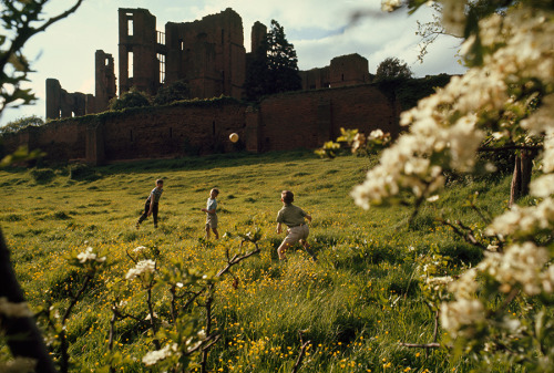 Young boys throw a ball on a lush green hillside below castle ruins in Warwickshire, England, 1968.P