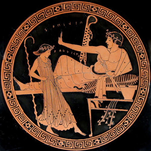 A youth reclines at a symposium, holding an aulos (double flute), while a woman dances in front of h