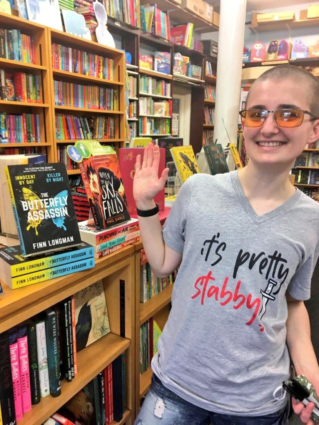 Finn Longman (a white person with a buzz cut and orange tinted glasses) wearing a grey t-shirt that reads "it's pretty stabby", with a picture of a knife. They're standing in a bookshop, gesturing towards copies of The Butterfly Assassin on display. 