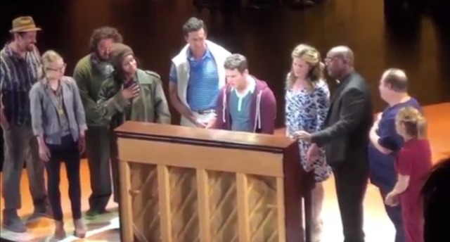 A screenshot from a bootleg of the 2015 version of the musical "A New Brain". It is taken during a scene where the main character, Gordon, (played by Jonathon Groff) sings and plays a song called "I Feel So Much Spring". The rest of the cast also stand around him on stage, singing along. 