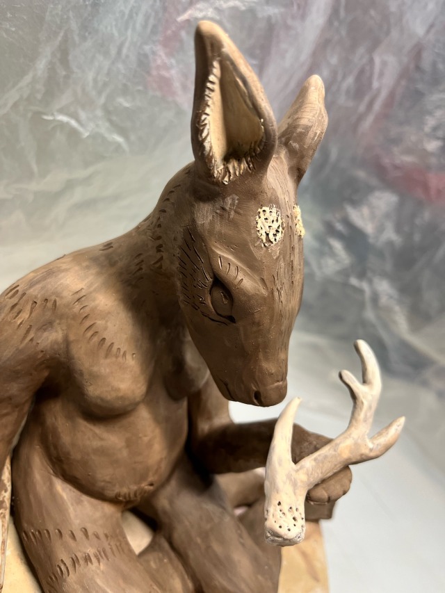 a brown clay sculpture of a sitting humanoid figure with the head of a deer. it\'s holding an antler and looking down at it thoughtfully