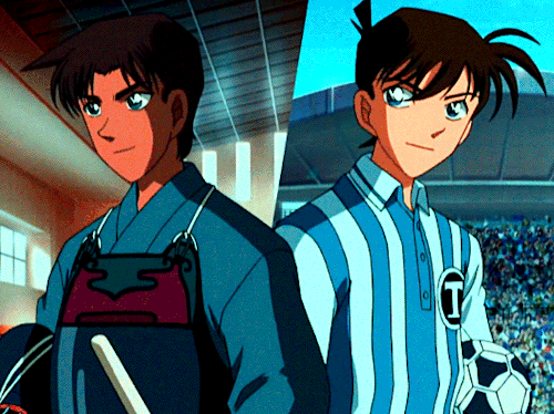 A GIF from the 15th Detective Conan/Case Closed opening sequence, which is set to the song "Hoshi no Kagayaki yo" by ZARD. Shinichi and Heiji are shown wearing sports' uniforms; Shinichi wears a soccer uniform for Teitan High School and holds a soccer ball, while Heiji wears a kendo uniform and holds his helmet. There's a line separating the two, with Shinichi at an outdoor soccer stadium and Heiji in an indoor gym. The camera then turns, and the two shift into wearing different outfits; Shinichi wears a blue suit with a red bow tie, while Heiji wears a blue jacket and his hat. They press their backs together and smile with their respective cityscapes behind them (with the landmark Osaka tower, Tsutenkaku, behind Heiji).
