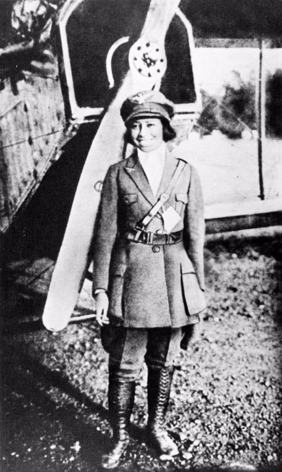 Black and white photograph of Bessie Coleman in a pilot attire, standing in front of an airplane.