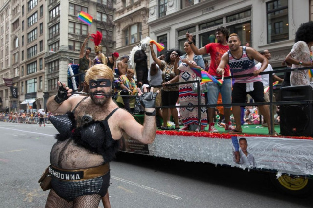 "Beardonna" a bearded man in costume with belly exposed and pointed leather brassiere bites a riding crop while behind a gay pride float rolls by at NYC pride event 
